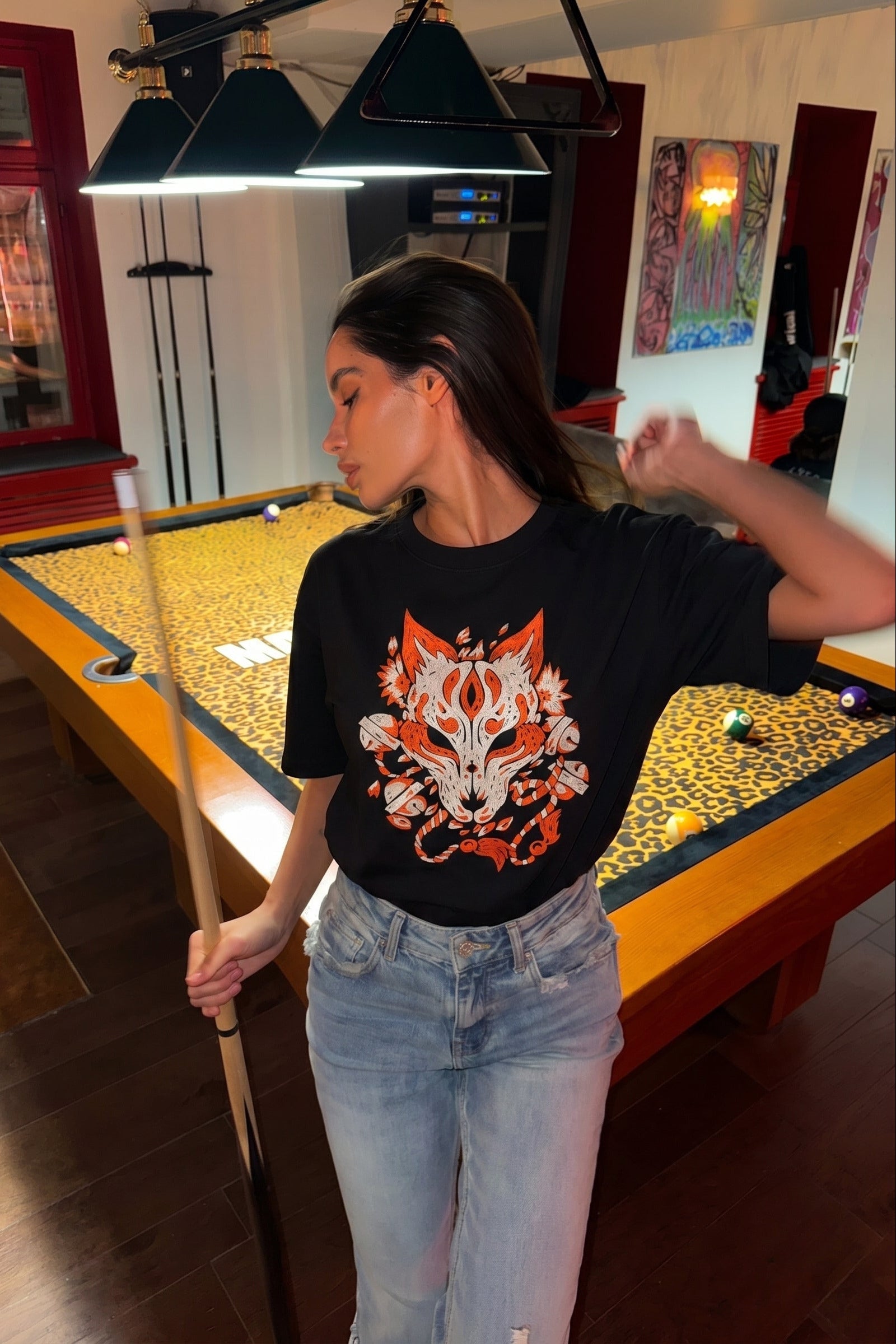 Woman in a black t-shirt with a fox design standing next to a pool table in a room with a pool table.