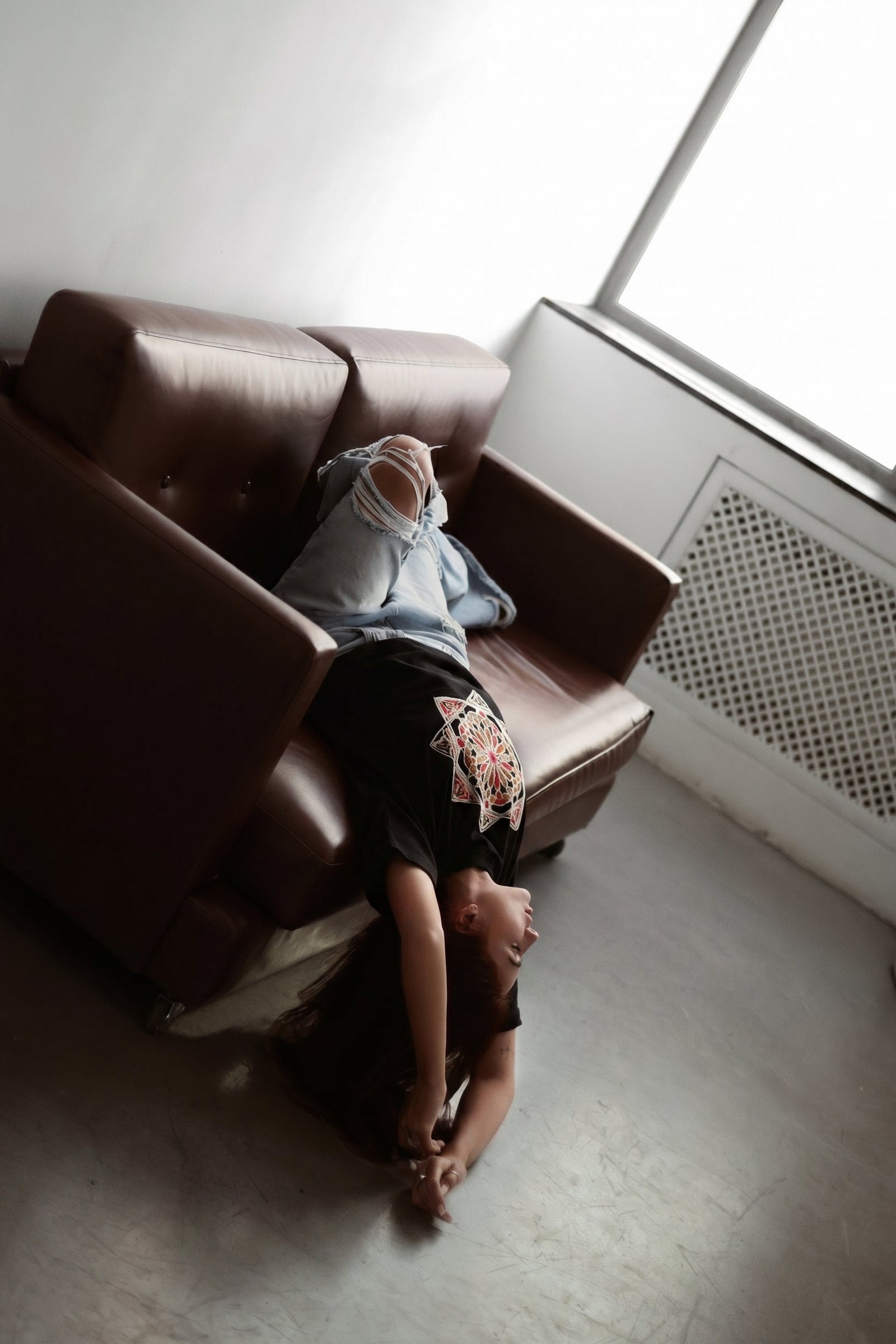 Person lying on a brown sofa in a room with a window and radiator.