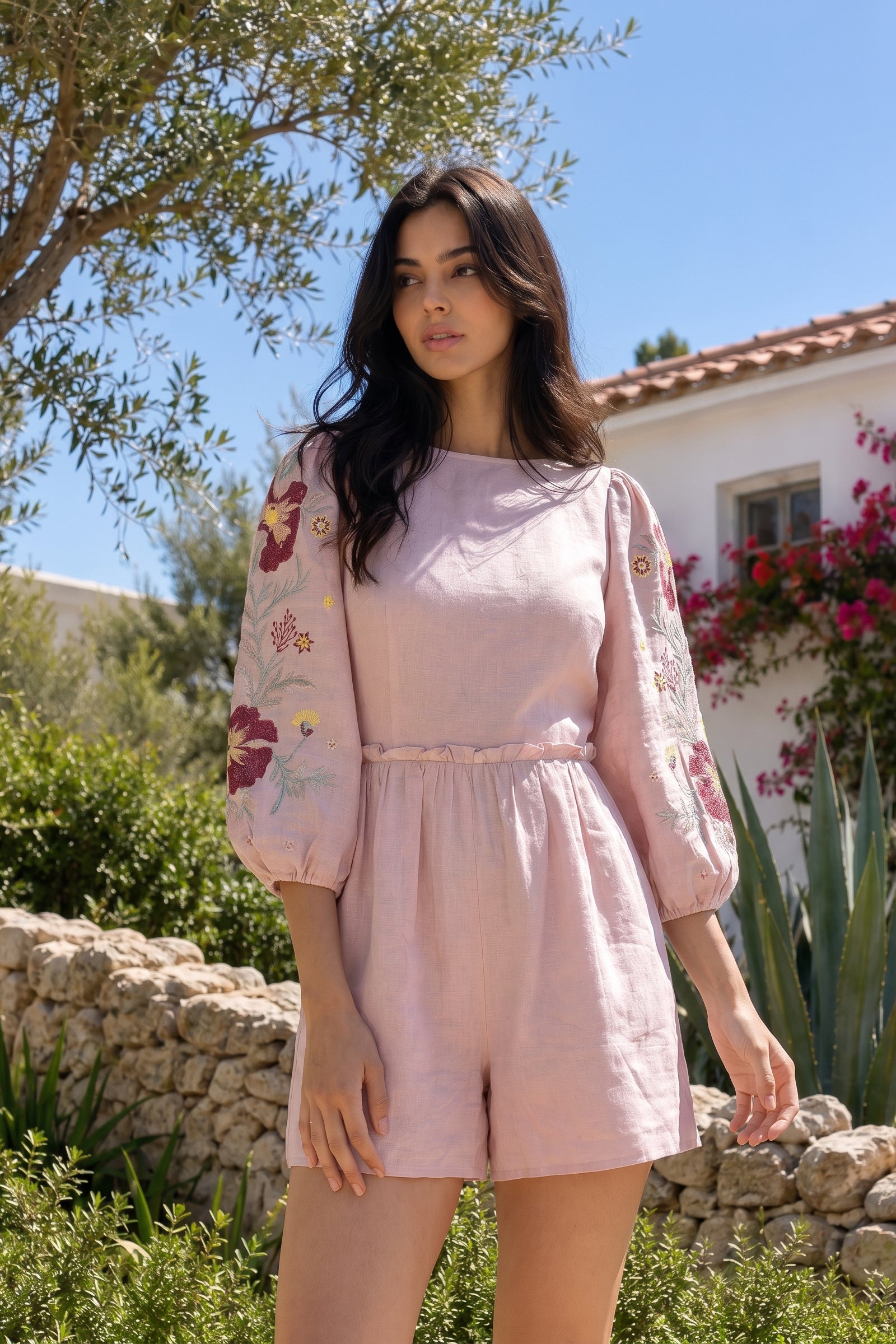 Woman in a pink outfit with floral embroidery standing outdoors near a stone wall and house.