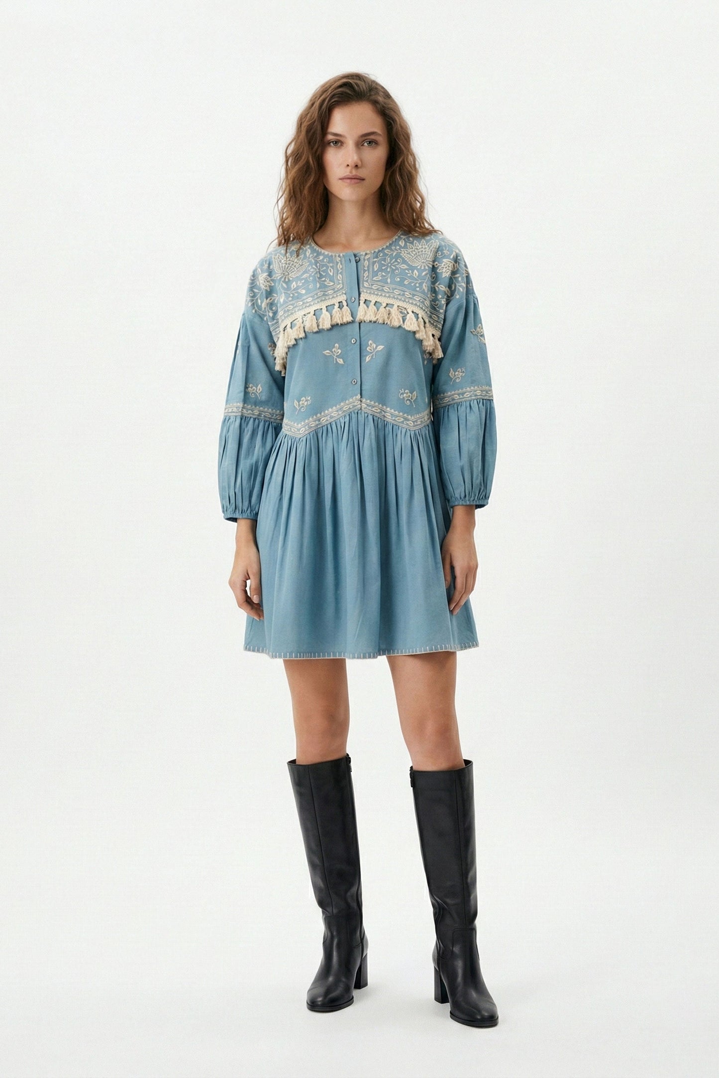 Woman wearing a blue embroidered dress with black boots on a white background