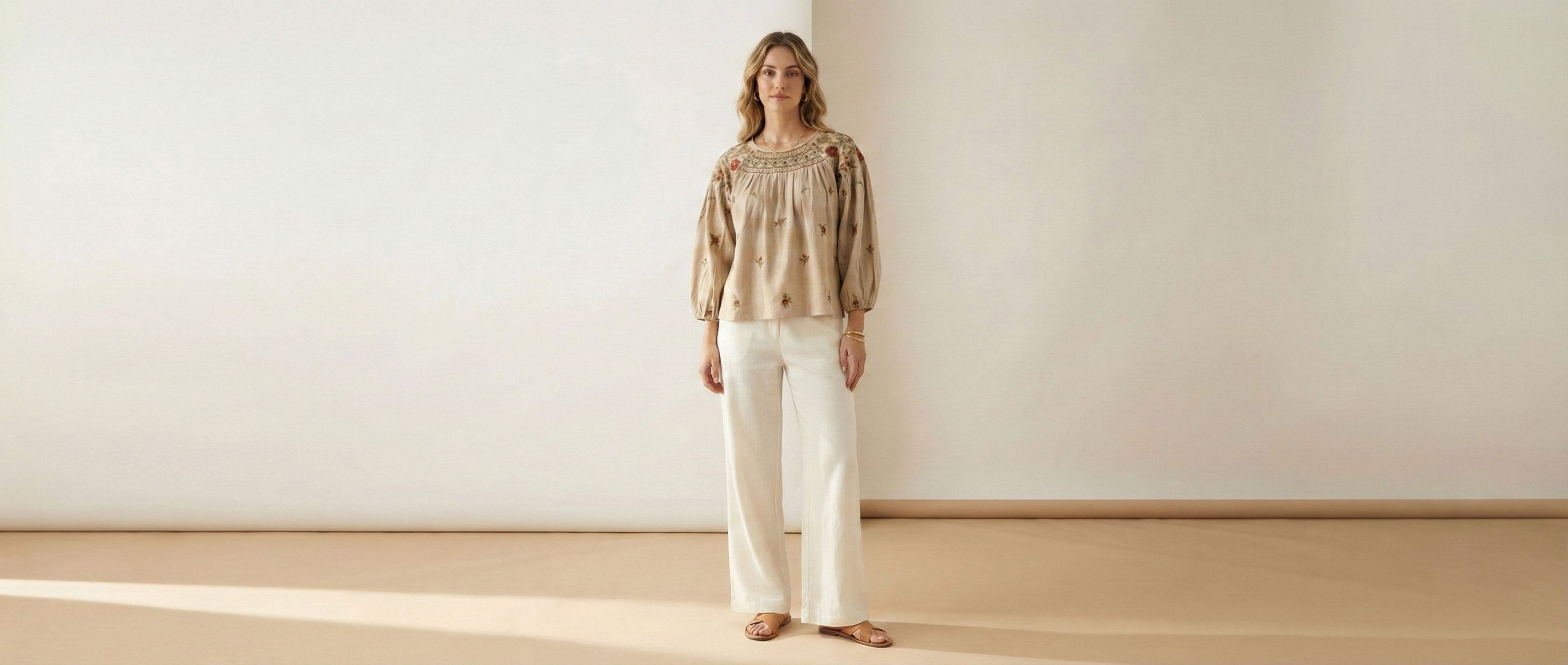 Woman wearing a patterned top and white pants standing in a minimalistic room with a white wall and wooden floor.