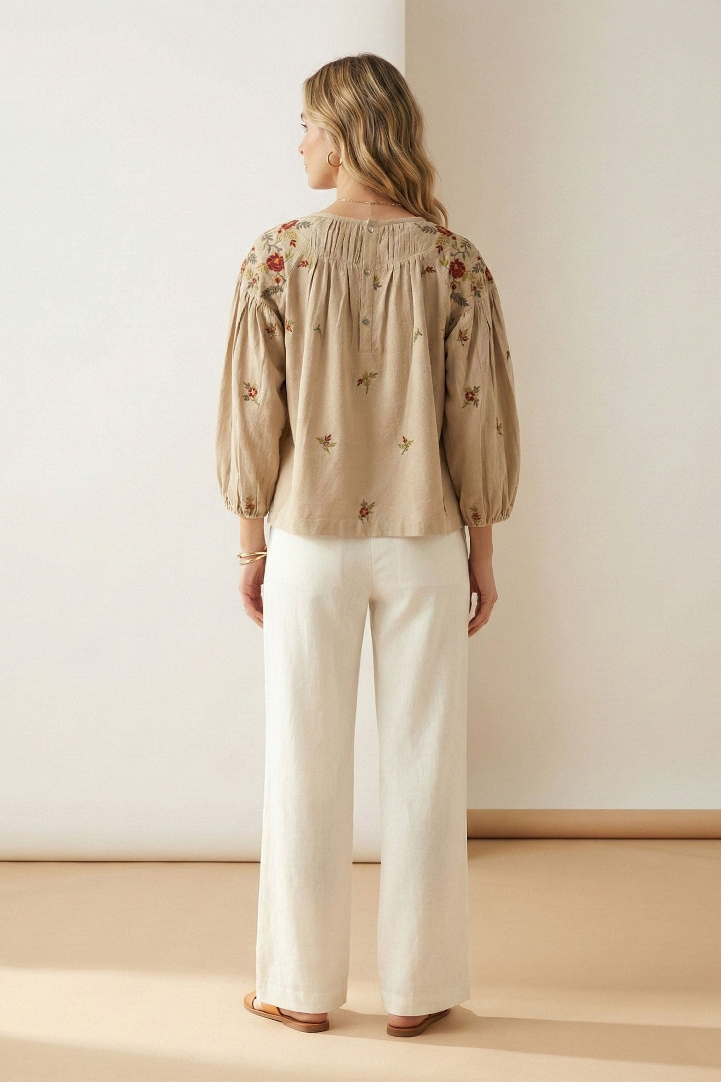 Woman wearing a beige embroidered blouse and white pants in a minimalistic setting