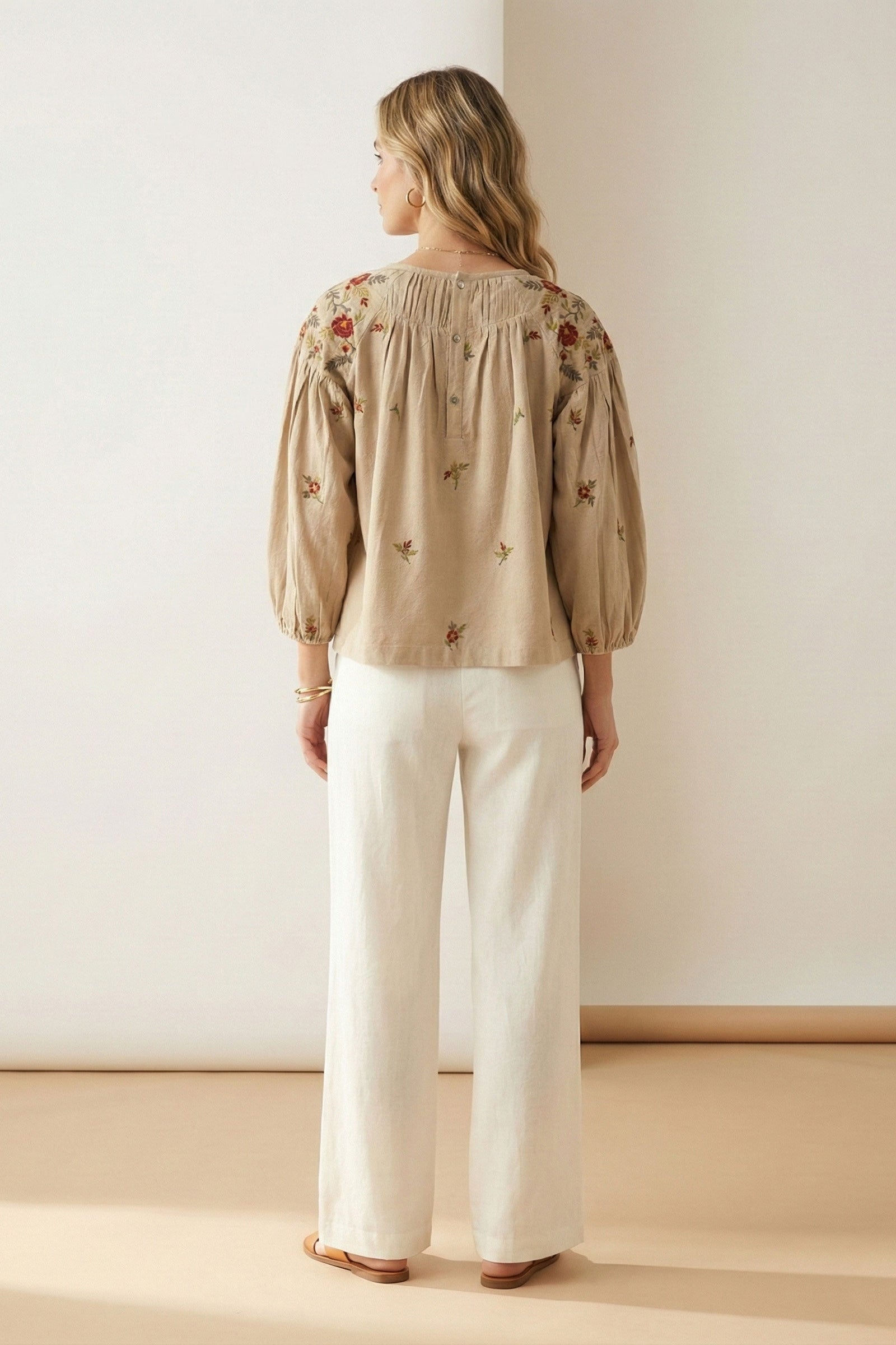 Woman wearing a beige embroidered blouse and white pants in a minimalistic setting