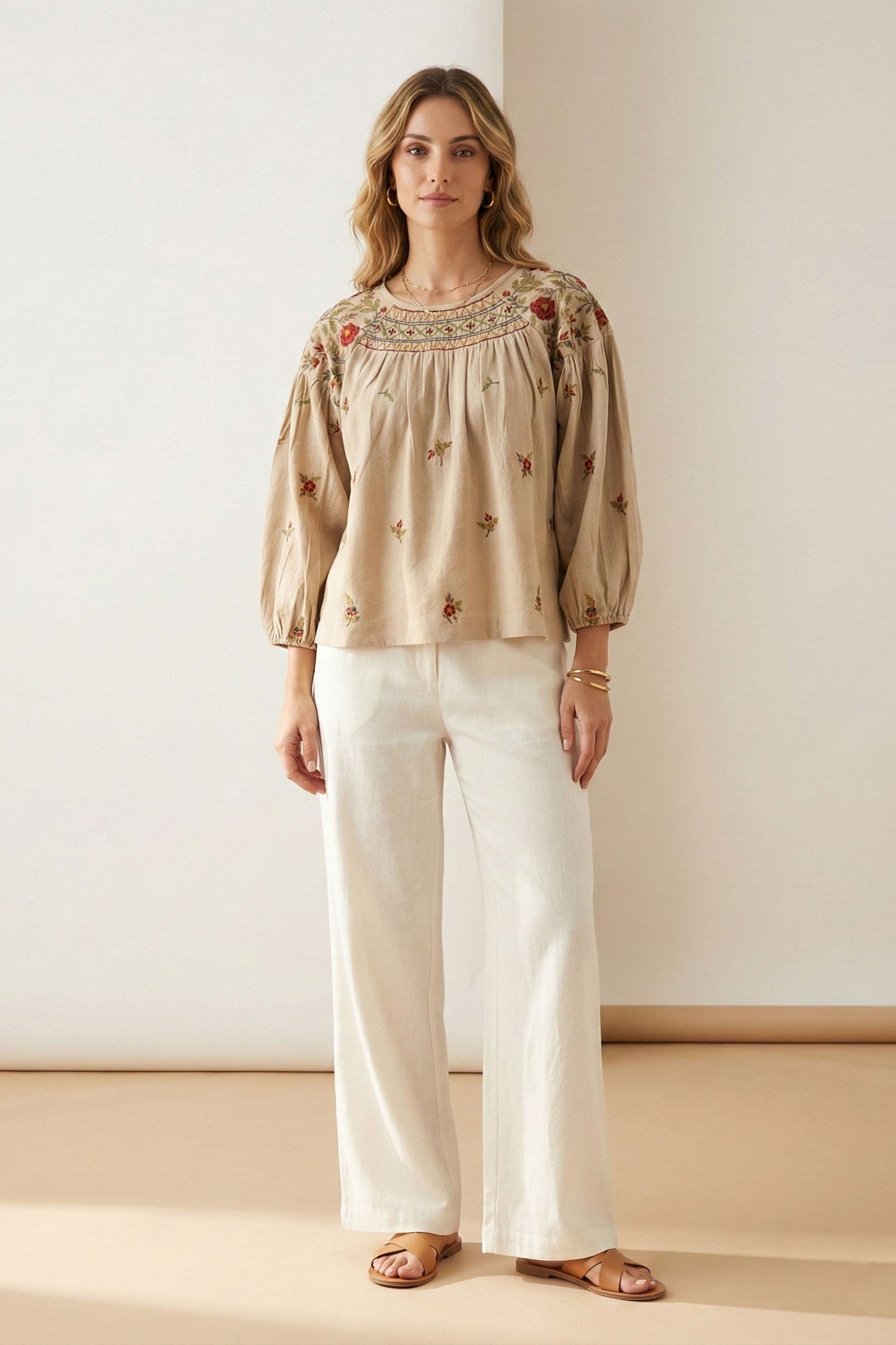 Woman wearing a beige embroidered blouse and white pants against a plain background
