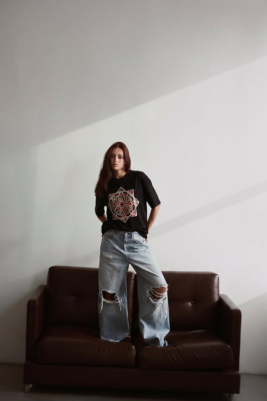 Person wearing a black t-shirt with a graphic design and light blue jeans standing on a brown couch against a white wall.