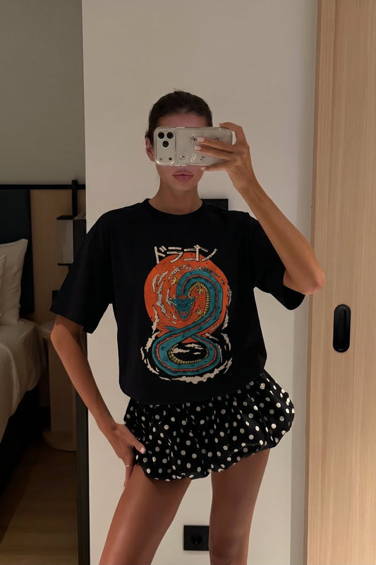 Person taking a mirror selfie wearing a black t-shirt with a colorful design and a polka dot skirt.